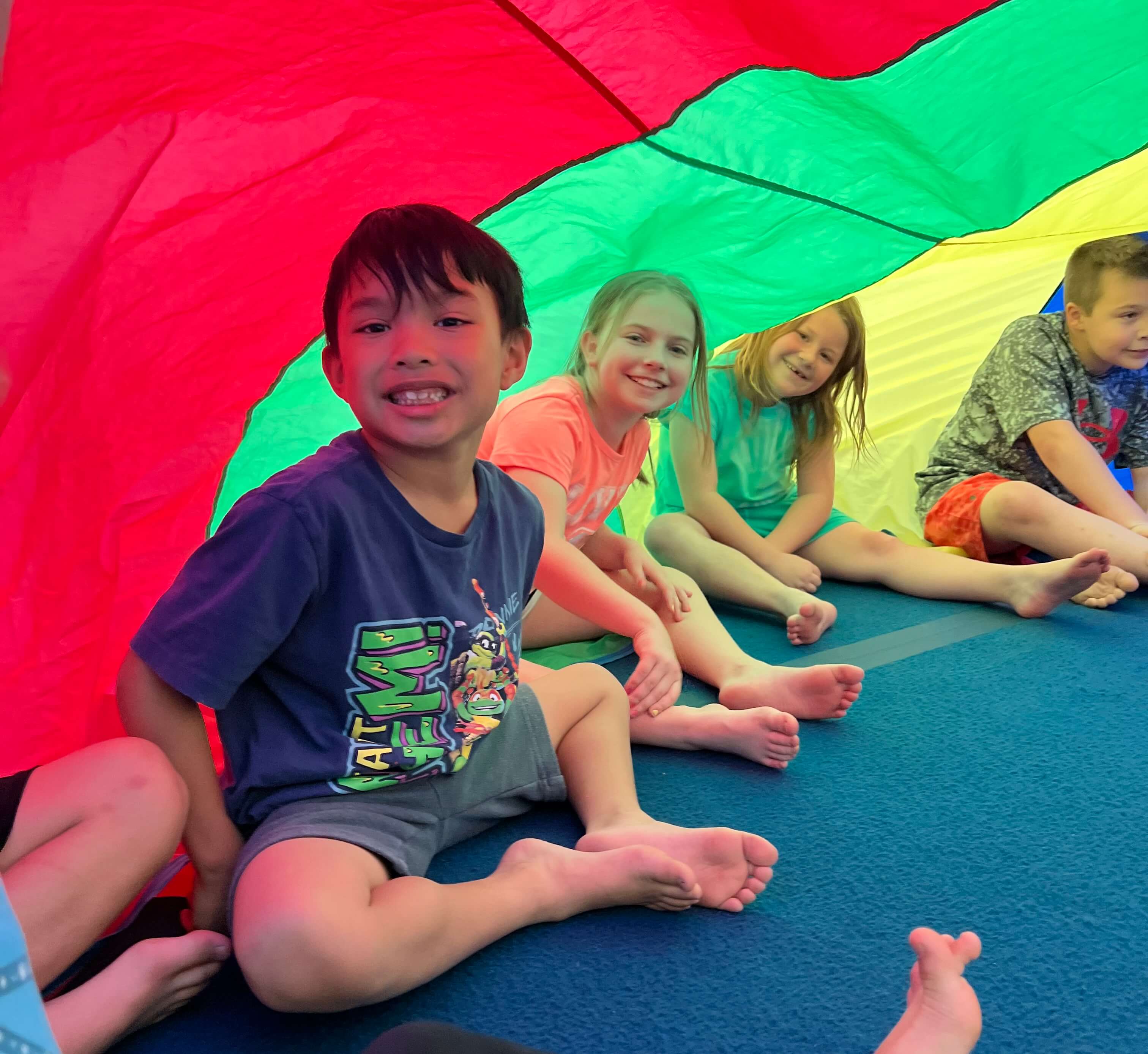 Kids sitting under a rainbow parachute at a gym party